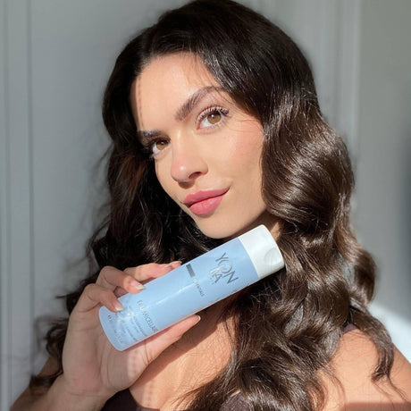 Young woman with long dark wavy hair holding a light blue bottle of Eau Micellaire Cleanser Makeup remover | Yon-Ka Paris in natural light, showcasing clear, glowing skin and promoting skincare wellness.