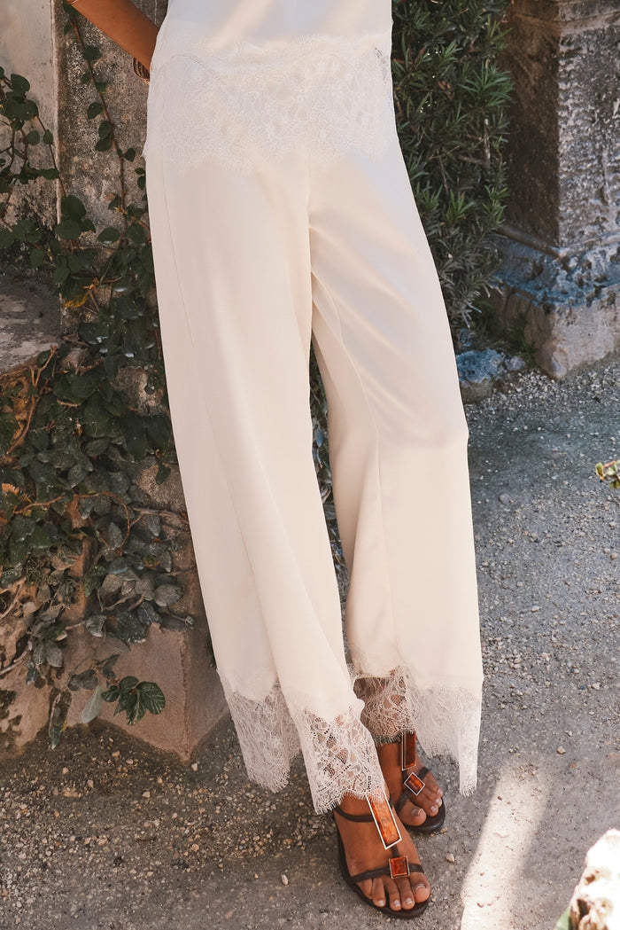 Cream satin lace-trim pants worn with tortoise sandals on a sunlit stone path.
