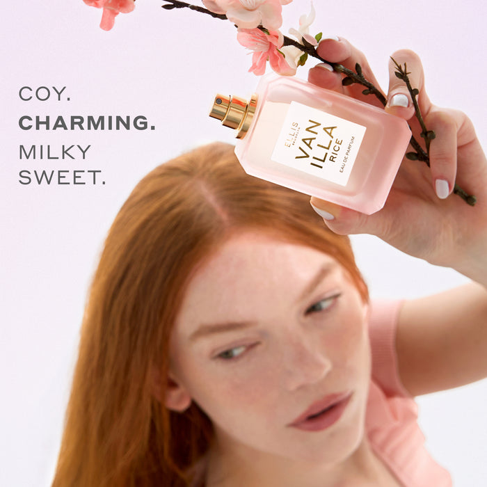 Woman with red hair wearing a pink shirt holds VANILLA RICE Eau de Parfum 50ml without cap and pink sakura blossoms above her head on a pink background with gray text that reads "coy. charming. milky sweet."